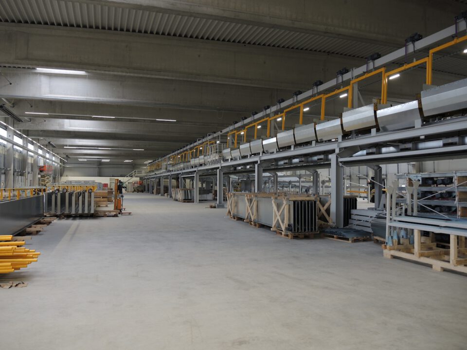 Innenansicht einer großen Industriehalle oder eines Lagers mit hohen Decken und heller Beleuchtung, in der lange Reihen von Maschinen und Förderbändern parallel verlaufen, umgeben von auf Paletten und in Kisten gelagerten Materialien und Bauteilen auf einem sauberen, weitläufigen Boden.