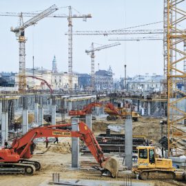 Das Bild zeigt eine große Baustelle mit mehreren Kränen und schweren Baumaschinen. Im Vordergrund arbeiten zwei Bagger, einer rot, der andere gelb, umgeben von Betonpfeilern und Trägern inmitten eines städtischen Umfelds.