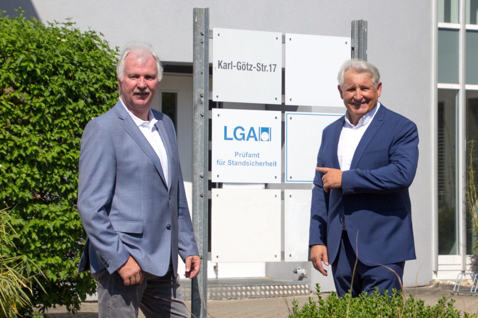 Zwei Männer stehen vor einem Schild mit mehreren Tafeln; oben steht „Karl-Götz-Str. 17“, darunter „LGA Prüfamt für Standsicherheit“, einer der Männer zeigt auf das Schild.