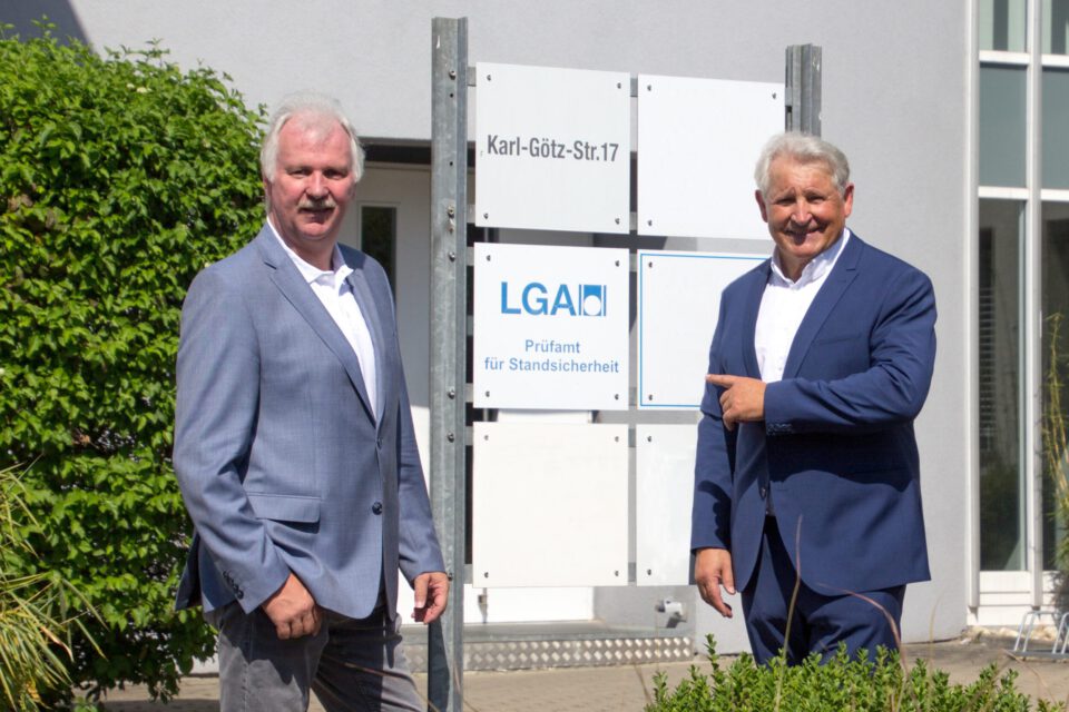 Zwei Männer stehen vor einem Schild mit mehreren Tafeln; oben steht „Karl-Götz-Str. 17“, darunter „LGA Prüfamt für Standsicherheit“, einer der Männer zeigt auf das Schild.