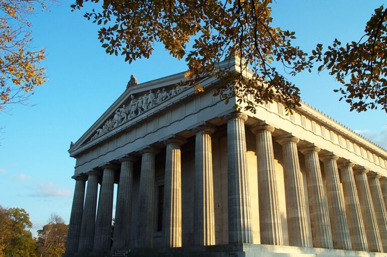 Das Bild zeigt ein klassizistisches Gebäude mit hohen Säulen und einem skulpturengeschmückten Giebelfeld, das an den Parthenon erinnert, bei Tageslicht und mit Bäumen im Vordergrund.