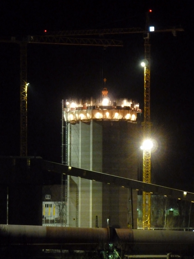 Nächtliche Baustelle mit zwei großen Kränen und einem beleuchteten hohen Bauwerk im Entstehungsprozess.