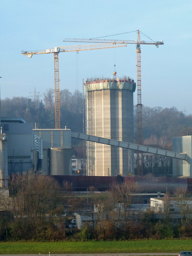 Baustelle mit zwei großen Kränen und einem hohen zylindrischen Bauwerk im Zentrum, das von Gerüsten und Industriegebäuden umgeben ist, außerdem einem Förderband sowie Bäumen und Hügeln im Hintergrund.