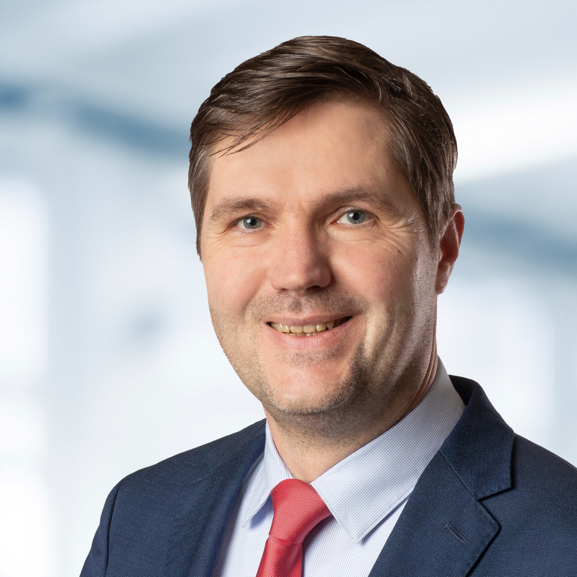 Dr. Marcus Achenbach trägt einen Anzug mit roter Krawatte. Der Hintergrund ist unscharf und zeigt ein Büro.