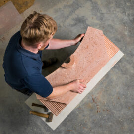 Eine Person arbeitet mit einem großen Stück Naturstein, das eine gebogene Form und eine strukturierte Oberfläche hat. Auf dem Boden liegen Werkzeuge wie Hammer und Meißel. Die Szene deutet auf handwerkliche Bearbeitung von Naturstein für Bauzwecke hin.
