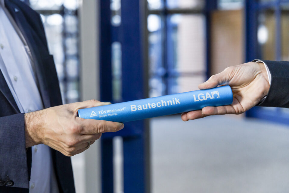 Das Bild zeigt zwei Personen, die sich einen blauen Staffelstab übergeben. Auf dem Stab steht „TÜV Rheinland Bautechnik LGA“. Die Szene symbolisiert eine Übergabe oder einen Wechsel.