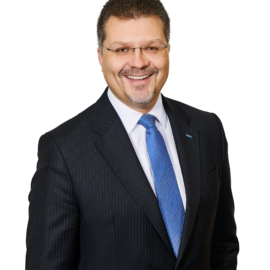 Das Bild zeigt eine Person in einem dunklen Nadelstreifenanzug mit weißem Hemd und blauer Krawatte. Der Hintergrund ist neutral und lenkt nicht vom Motiv ab.