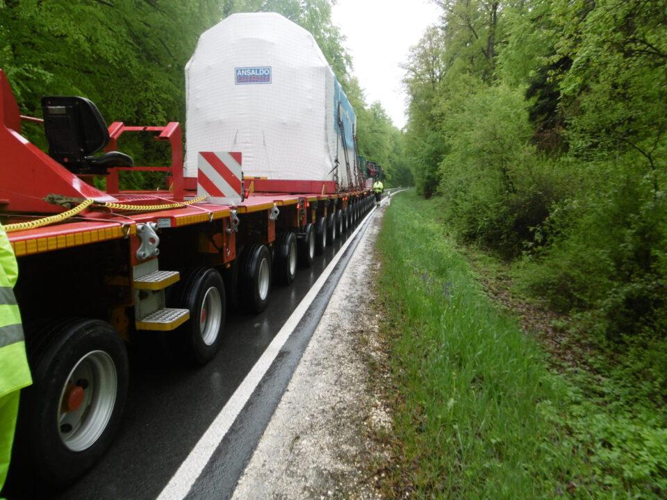Das Bild zeigt ein großes Transportfahrzeug mit mehreren Achsen, das ein riesiges, verpacktes Objekt auf einer schmalen Straße transportiert. Die Umgebung ist von dichtem, grünem Baumbestand gesäumt.