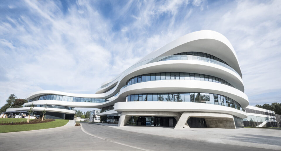Das Bild zeigt ein modernes, weißes Gebäude mit geschwungener Architektur und großen Glasflächen. Die Form des Gebäudes erinnert an die Struktur von Wellpappe, passend zum Produkt des Unternehmens. Es handelt sich um ein multifunktionales Firmengebäude mit nachhaltigen Bau- und Energiekonzepten.
