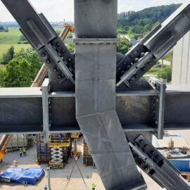 Das Bild zeigt eine Nahaufnahme einer großen Stahlkonstruktion mit mehreren sich kreuzenden Stahlträgern, die durch Schrauben und Platten verbunden sind. Im Hintergrund ist eine Baustelle mit Kränen, Geräten und Arbeitern zu sehen. Die Umgebung besteht aus grünen Feldern und Bäumen, was auf eine ländliche oder halbländliche Lage hinweist.
