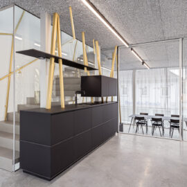 Das Bild zeigt einen modernen, minimalistisch gestalteten Innenraum mit einer Treppe auf der linken Seite. Die Treppe besteht aus Metallstufen, einem Glasgeländer und goldfarbenen Handläufen. Daneben steht ein schwarzes Sideboard mit Schubladen und vertikalen Holzelementen, im Hintergrund trennt eine Glaswand den Raum von einem weiteren Bereich mit Tisch und Stühlen.