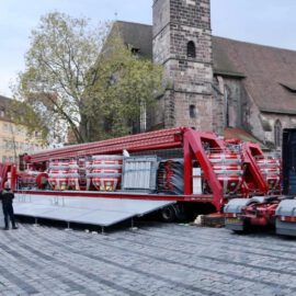 Das zweite Bild zeigt den Aufbau eines großen rot-weißen Fahrgeschäfts auf einem gepflasterten Platz in einer Stadt. Das Fahrgeschäft befindet sich noch auf einem Anhänger, der an einen LKW gekoppelt ist. Mehrere Personen arbeiten rund um das Fahrgeschäft, vermutlich beim Aufbau. Im Hintergrund sind Gebäude und eine Kirche zu sehen, was auf eine zentrale Lage – möglicherweise ein Marktplatz – hinweist.