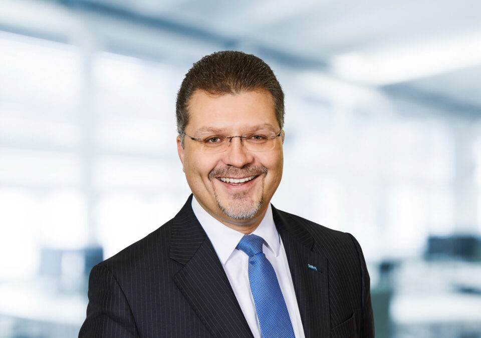 Das Bild zeigt eine Person in einem dunklen Nadelstreifenanzug mit weißem Hemd und blauer Krawatte. Der Hintergrund wirkt wie ein Büro mit unscharfen Fenstern und Möbeln.