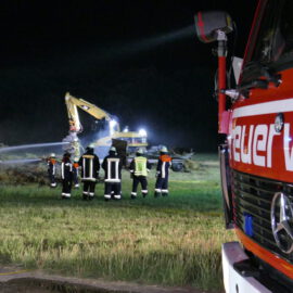 Das Bild zeigt eine nächtliche Einsatzszene der Feuerwehr auf einem grasbewachsenen Feld. Im Vordergrund steht ein rotes Feuerwehrfahrzeug, mehrere Feuerwehrleute in Schutzkleidung sind im Einsatz. Zwei Feuerwehrleute sprühen Wasser, während helle Scheinwerfer die Umgebung ausleuchten.