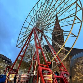Ein großes Riesenrad wird in einer städtischen Umgebung aufgebaut oder abgebaut. Die Metallstruktur ist teilweise errichtet und wird von roten Trägern gestützt, während mehrere Personen am Boden arbeiten. Im Hintergrund sind eine Kirche mit hohem Turm und weitere Gebäude zu sehen, der Himmel ist in einem blauen Ton gehalten.