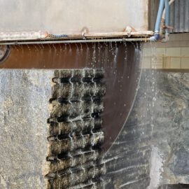 Das Bild zeigt ein großes industrielles Sägeblatt, das einen Steinblock durchtrennt. Wasser wird auf das Sägeblatt und den Stein gesprüht, um das Blatt zu kühlen und Staub zu reduzieren. Der Steinblock weist mehrere horizontale Rillen auf, die auf eine Teilung in kleinere Abschnitte hinweisen.