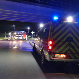 Das Bild zeigt eine nächtliche Einsatzszene unter einer Brücke mit einem Feuerwehrfahrzeug und einem weiteren Einsatzfahrzeug mit eingeschaltetem Blaulicht. Auf der Straße liegen Schläuche, und mehrere Einsatzkräfte sind im Arbeitsbereich zu sehen. Die Szene wird von Straßenlaternen beleuchtet.