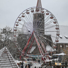 Das Bild zeigt einen verschneiten Stadtplatz mit einem großen, beleuchteten Riesenrad in der Mitte. Im Hintergrund steht eine Kirche mit spitzem Turm, und rund um das Riesenrad sind Menschen unterwegs, teils an kleinen Marktständen. Die Dächer der Gebäude sind ebenfalls mit Schnee bedeckt und unterstreichen die winterliche Stimmung.