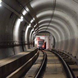 Das Bild zeigt einen U-Bahn-Tunnel, in dem ein Zug aus der Ferne heranfährt. Der Tunnel ist gut beleuchtet, mit einer begehbaren Fläche links neben den Gleisen und gebogenen Betonwänden.