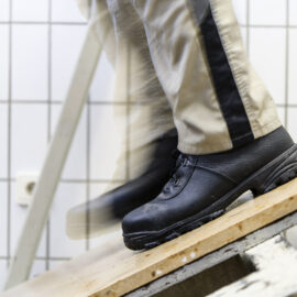 Das Bild zeigt eine Person mit schwarzen Arbeitsschuhen und beigefarbener Hose mit schwarzem Seitenstreifen, die über ein Holzbrett läuft. Der Hintergrund besteht aus weißen Fliesen mit einem Gittermuster. Eine Bewegungsunschärfe deutet auf eine laufende Bewegung hin.