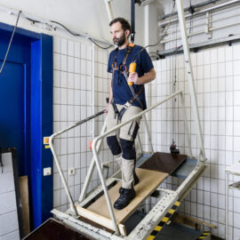 Das Bild zeigt eine Person, die auf einem schräg gestellten Hebebühne steht, das Teil eines Labors oder Testaufbaus zu sein scheint. Die Person ist mit Sicherheitsgurten an einem Rahmen über dem Gerät gesichert. Im Hintergrund sind geflieste Wände, Kabel und technische Geräte zu sehen, die auf eine kontrollierte Versuchsumgebung hinweisen.