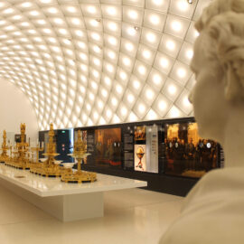 Das Bild zeigt den Innenraum eines Museums oder einer Ausstellungshalle mit moderner Deckenbeleuchtung. Im Vordergrund steht eine weiße Marmorbüste, in der Mitte ein langer weißer Ausstellungstisch mit goldenen Objekten und Skulpturen. An den Wänden befinden sich ein gerahmtes Porträtgemälde sowie Glasvitrinen mit weiteren Exponaten und Informationstafeln.