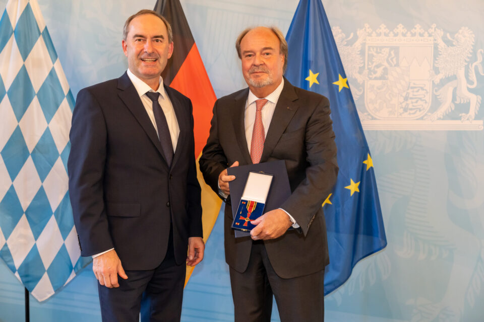 Zwei Männer im Anzug stehen nebeneinander. Der Mann rechts hält eine geöffnete Schachtel mit einer Medaille oder Auszeichnung. Im Hintergrund sind die Flaggen von Bayern, Deutschland und der Europäischen Union zu sehen.