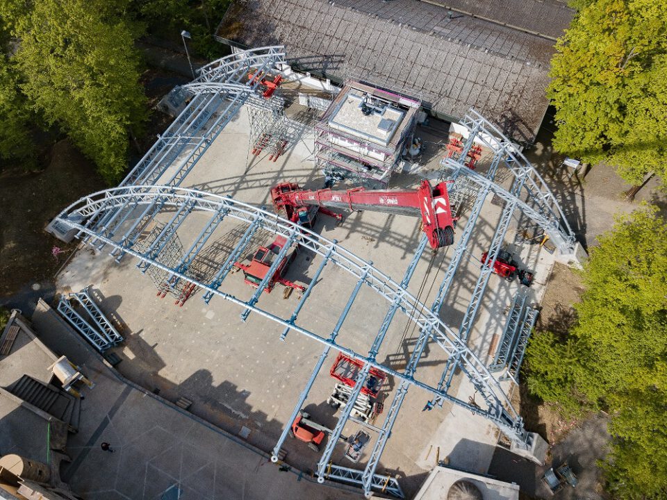 Das Bild zeigt eine Luftaufnahme einer Baustelle mit einer großen Metallkonstruktion, die das Gerüst eines Gebäudes oder Pavillons bildet. Innerhalb der Struktur befinden sich mehrere rote Baustellenfahrzeuge wie Kräne und Hebebühnen. Im Hintergrund sind Bäume und angrenzende Gebäude zu sehen.