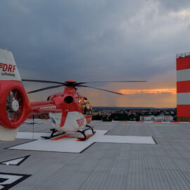 Das Bild zeigt einen rot-weißen Rettungshubschrauber auf einem Landeplatz. Im Hintergrund ist eine Stadtlandschaft bei Sonnenuntergang zu sehen, mit Wolken am Himmel und einem hohen rot-weiß gestreiften Turm.