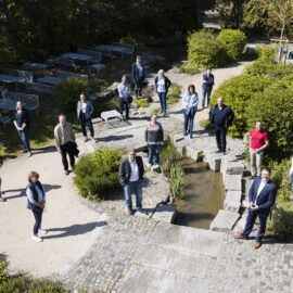 Das Bild zeigt eine Gruppe von Menschen, die im Freien in einer gartenähnlichen Umgebung stehen und Abstand zueinander halten. In der Mitte befindet sich ein kleines Wasserbecken mit Trittsteinen, umgeben von Pflanzen und Sträuchern. Im Hintergrund sind Tische, Bänke und ein Gebäude auf der rechten Seite zu sehen.