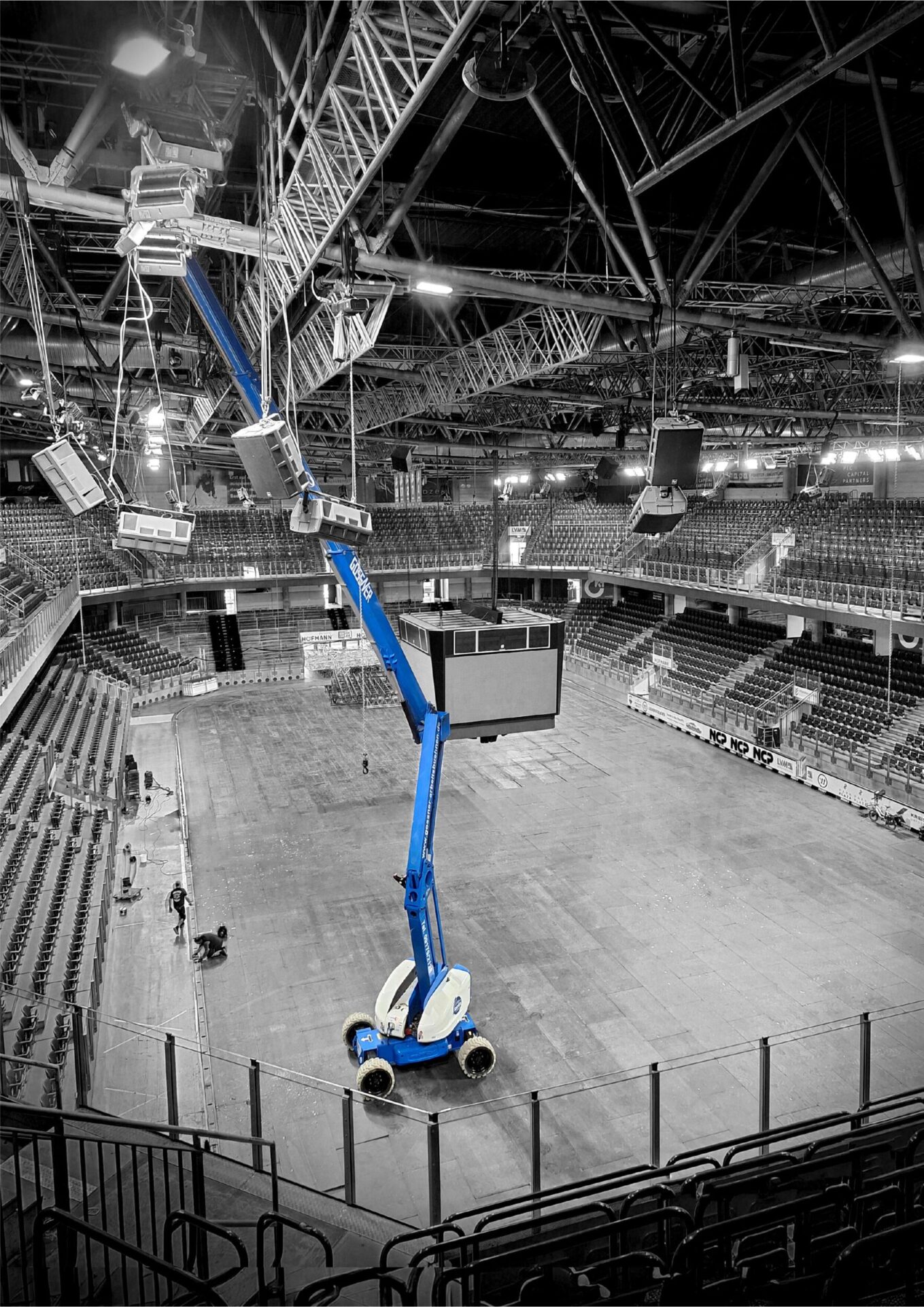 In der Aufnahme ist das Innere einer großen Veranstaltungshalle zu sehen, mit vielen Sitzreihen rund um eine freie Fläche in der Mitte. Ein blauer Gelenkteleskoparbeitsbühnenlift steht auf dem Boden und reicht mit seinem langen Arm bis zur Decke, wo er an der Beleuchtung oder Technik arbeitet. Über dem Lift sind zahlreiche Metallträger, Lautsprecher und Beleuchtungselemente an der Hallendecke angebracht.