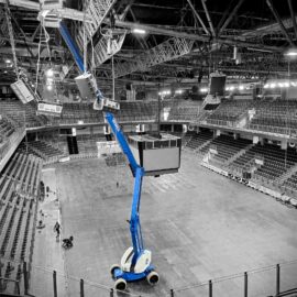 In der Aufnahme ist das Innere einer großen Veranstaltungshalle zu sehen, mit vielen Sitzreihen rund um eine freie Fläche in der Mitte. Ein blauer Gelenkteleskoparbeitsbühnenlift steht auf dem Boden und reicht mit seinem langen Arm bis zur Decke, wo er an der Beleuchtung oder Technik arbeitet. Über dem Lift sind zahlreiche Metallträger, Lautsprecher und Beleuchtungselemente an der Hallendecke angebracht.