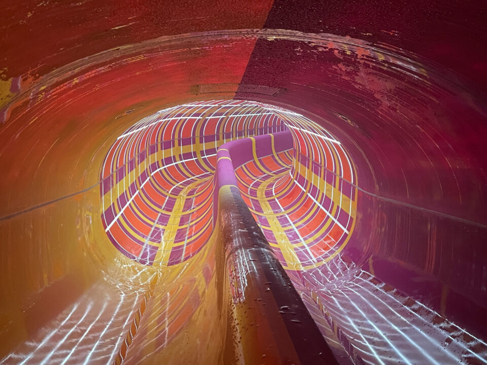 Das Bild zeigt das Innere einer bunten Wasserrutsche mit leuchtenden Streifen in Rot, Orange, Gelb und Lila. Die Oberfläche ist nass und glänzend, was auf eine Nutzung oder Betriebsbereitschaft hinweist.