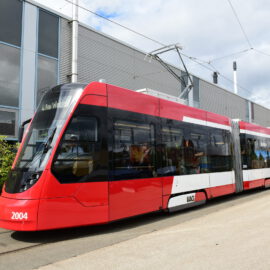Ein moderner rot-weißer Straßenbahnwagen mit der Nummer 2004 steht auf einem Gleis neben einem großen Industriegebäude mit grauer Verkleidung und mehreren Fenstern. Die Straßenbahn ist über Oberleitungen elektrisch angeschlossen und zeigt die Liniennummer 4 mit dem Ziel „Am Wegfeld“. Die Umgebung wirkt wie ein Industrie- oder Wartungsbereich.