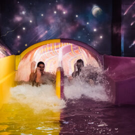 Das Bild zeigt zwei Personen, die nebeneinander eine Wasserrutsche hinunter in ein Becken rutschen. Die Rutsche ist zweigeteilt mit einer gelben und einer lilafarbenen Bahn. Im Hintergrund ist ein Wandbild mit Weltraummotiven wie Sternen und Planeten zu sehen.