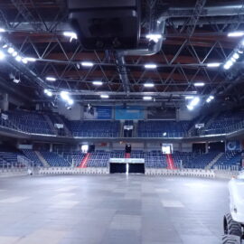 Die Aufnahme zeigt das Innere einer großen Mehrzweckhalle mit leeren Sitzreihen, die sich über mehrere Ebenen erstrecken. In der Mitte befindet sich eine freie Fläche ohne Bestuhlung, während an der Decke zahlreiche Lichtstrahler und Metallträger sichtbar sind. Am rechten Bildrand steht ein weißes Arbeitsbühnenfahrzeug, das teilweise im Bild zu sehen ist.