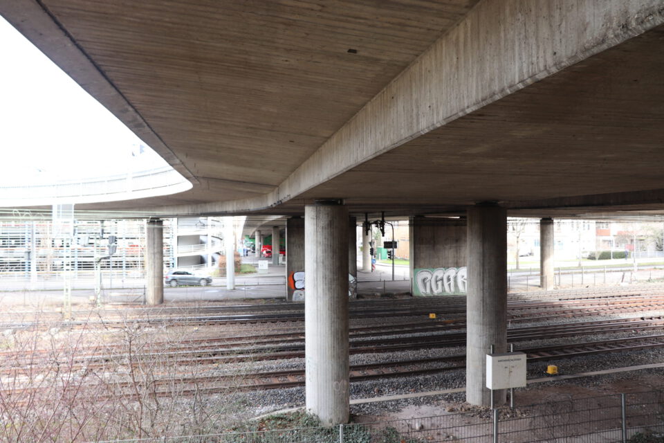 Das Bild zeigt eine Unterführung mit mehreren Betonpfeilern, die eine große Überbauung wie eine Brücke oder Autobahn tragen. Unter der Konstruktion verlaufen mehrere parallel angeordnete Bahngleise. Im Hintergrund ist Graffiti auf einem der Pfeiler zu sehen, während im Vordergrund etwas Vegetation und im Hintergrund städtische Gebäude erkennbar sind.