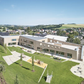 Das Bild zeigt ein modernes, zweigeschossiges Gebäude mit Flachdach in einer grünen, gestalteten Umgebung. Vor dem Gebäude befinden sich Spielgeräte und neu gepflanzte Bäume. Im Hintergrund sind Wohnhäuser und sanfte Hügel unter einem klaren blauen Himmel zu sehen.