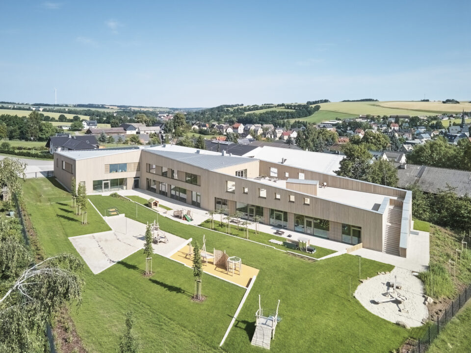 Das Bild zeigt ein modernes, zweigeschossiges Gebäude mit Flachdach in einer grünen, gestalteten Umgebung. Vor dem Gebäude befinden sich Spielgeräte und neu gepflanzte Bäume. Im Hintergrund sind Wohnhäuser und sanfte Hügel unter einem klaren blauen Himmel zu sehen.