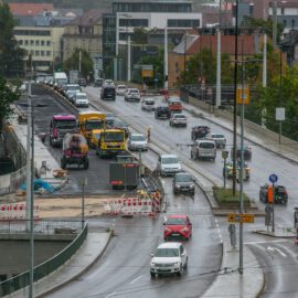 Das Bild zeigt eine stark befahrene städtische Straße mit mehreren Fahrspuren, auf denen Autos und Lastwagen unterwegs sind. Auf der linken Seite findet eine Straßenbaumaßnahme statt, erkennbar an Baustellenfahrzeugen und Absperrungen. Der nasse Straßenbelag und Lichtreflexionen deuten auf regnerisches Wetter hin, im Hintergrund sind Gebäude und Bäume sichtbar.