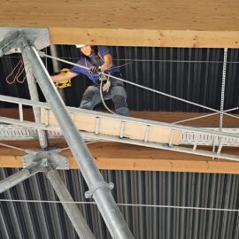 Das Bild zeigt eine Person, die auf einer schmalen Arbeitsplattform in großer Höhe arbeitet. Die Plattform ist zwischen Metallträgern und Holzbalken befestigt, und die Person trägt eine Sicherungsausrüstung mit Seilen. Im Hintergrund sind eine dunkle Metallverkleidung und Teile der Dachkonstruktion sichtbar.