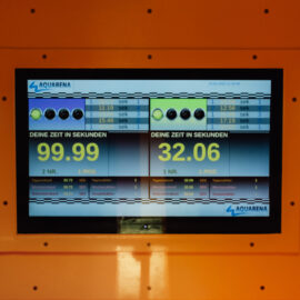 Das Bild zeigt einen Bildschirm mit mehreren Zahlenanzeigen und einem orangefarbenen Rahmen mit Knöpfen und Lautsprechern. Auf dem Display sind zwei Zeitmessungen in Sekunden sowie weitere kleinere Zeiten dargestellt. Der Bildschirm gehört zu einer Anlage zur Zeitmessung, vermutlich im Bereich Wasserrutschen.