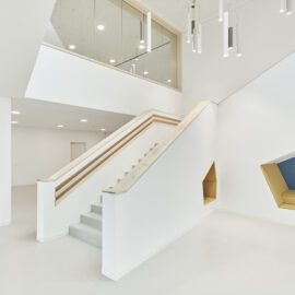 Das Bild zeigt einen modernen, hellen Innenraum mit weißen Wänden und Decken sowie einer Treppe mit Holzhandlauf, die zu einer oberen Ebene mit Glasbrüstung führt. An der Decke befinden sich eingelassene Leuchten und zylindrische Hängelampen. Rechts ist eine in die Wand integrierte Sitznische mit gelben Polstern und blauer Rückenlehne zu sehen.