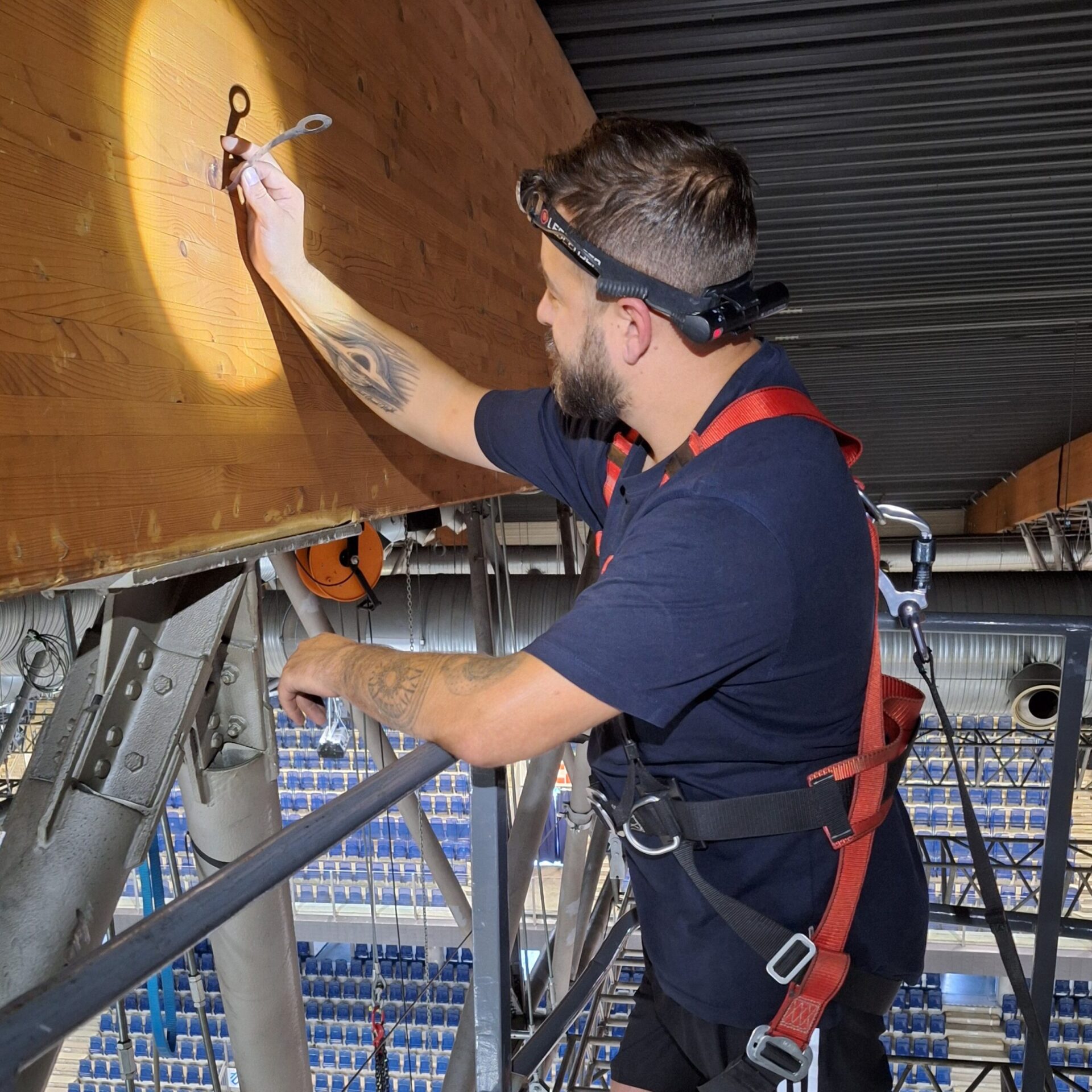 Das Bild zeigt eine Person, die auf einer Metallkonstruktion in großer Höhe arbeitet und dabei eine Sicherungsausrüstung trägt. Die Person befestigt ein Metallteil an einem großen Holzbalken, der Teil der Dachkonstruktion ist. Im Hintergrund sind Sitzreihen einer Halle sowie weitere Metallträger und technische Installationen sichtbar.