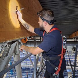 Das Bild zeigt eine Person, die auf einer Metallkonstruktion in großer Höhe arbeitet und dabei eine Sicherungsausrüstung trägt. Die Person befestigt ein Metallteil an einem großen Holzbalken, der Teil der Dachkonstruktion ist. Im Hintergrund sind Sitzreihen einer Halle sowie weitere Metallträger und technische Installationen sichtbar.