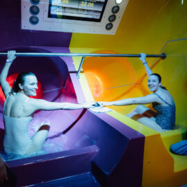 Das Bild zeigt zwei Personen, die am Einstieg zweier Wasserrutschen sitzen, eine in Lila und eine in Gelb. Sie reichen sich über eine Trennwand hinweg die Hände. Ein Mann steht daneben und beobachtet die Szene, über den Rutschen ist eine Anzeigetafel mit Zahlen zu sehen.