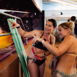 Zwei Personen in Badekleidung deuten nach links. Neben ihnen an ein Becken angelehnt 3 Schwimmnudeln. Im Hintergrund weitere Personen.