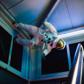 Das Bild zeigt einen Astronauten in einem weißen Raumanzug, der innerhalb einer Raumstation oder eines Raumschiffs schwebt. Der Astronaut ist waagerecht ausgerichtet, mit dem Kopf nach rechts und den Füßen nach links. Im Hintergrund sind metallische Wände und Fenster zu sehen.