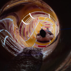 Das Bild zeigt eine Person, die eine geschlossene Wasserrutsche hinunterrutscht. Die Innenwände der Rutsche sind bunt gemustert und werden von hellen Lichtern beleuchtet. Die Person sitzt aufrecht und blickt in Fahrtrichtung.