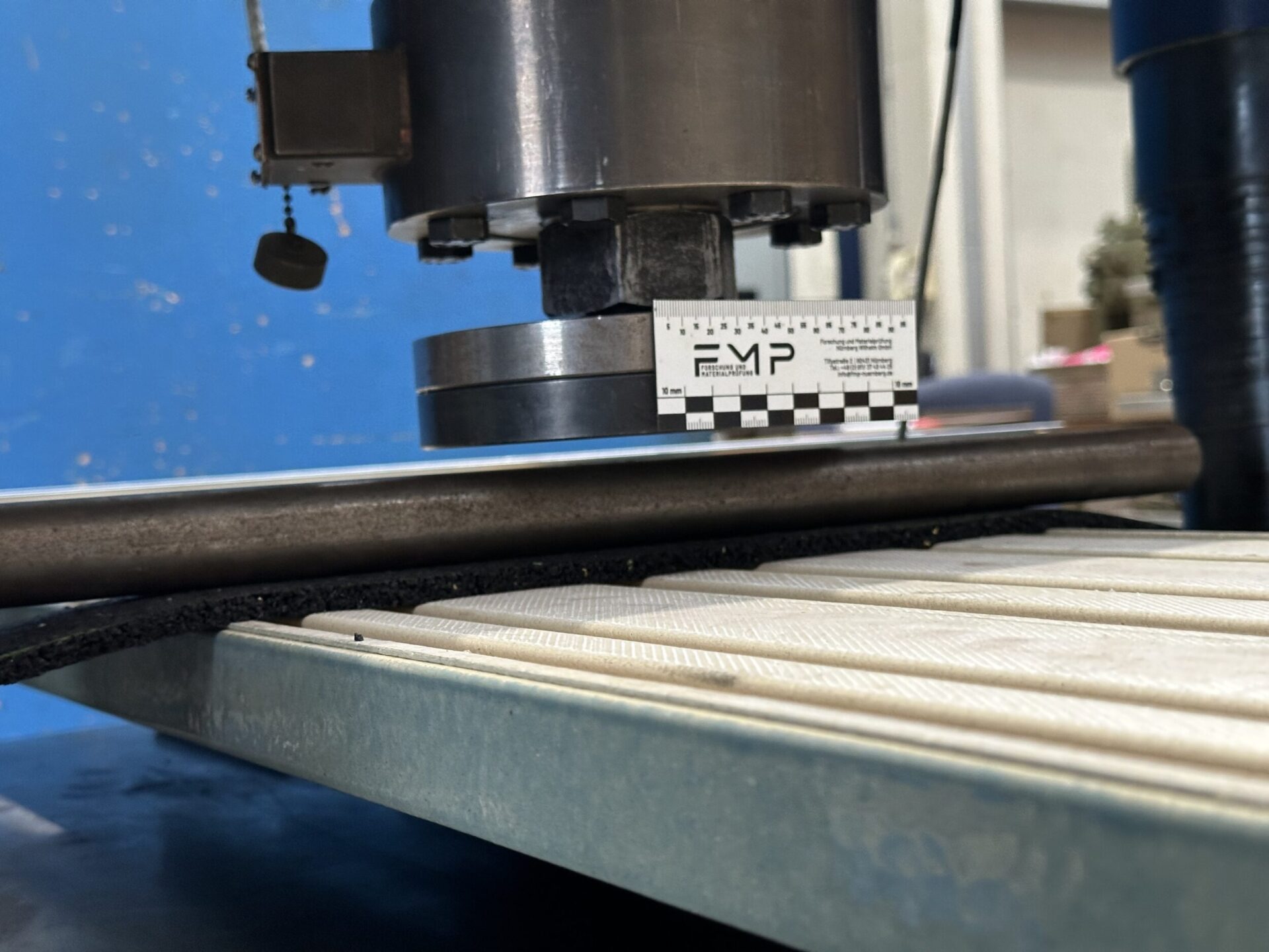 Das Bild zeigt eine Nahaufnahme einer Prüfmaschine in einem technischen Umfeld. Im Vordergrund ist ein Metallstab eingespannt, darüber befindet sich ein zylindrisches Prüfwerkzeug, das auf den Stab drückt. Daneben liegt ein Maßstab zur Größenreferenz, und im Hintergrund sind unscharfe Werkstattdetails zu erkennen.